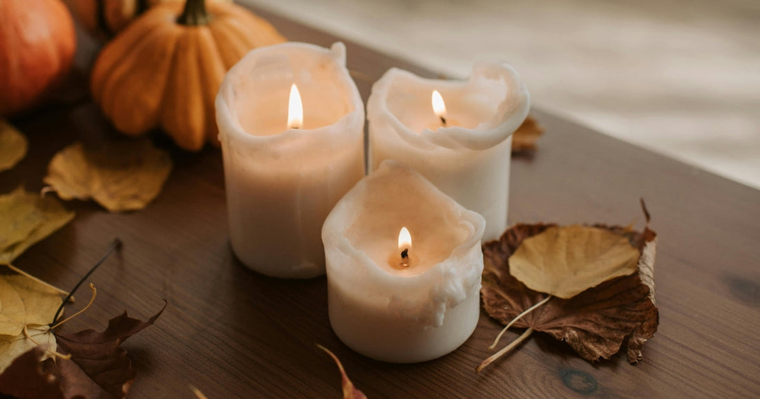 Three white candles burning near dried fall leaves & pumpkins, creating a cozy fall vibe.